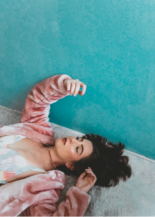 a person dressed in a furry pink jacket and a flowered dress lying on their back on a light grey carpet with a teal wall in the background