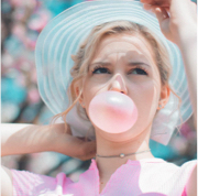 a whimsical headshot of a person blowing a pink bubble with their gum while wearing a matching pink shirt and a light blue hat