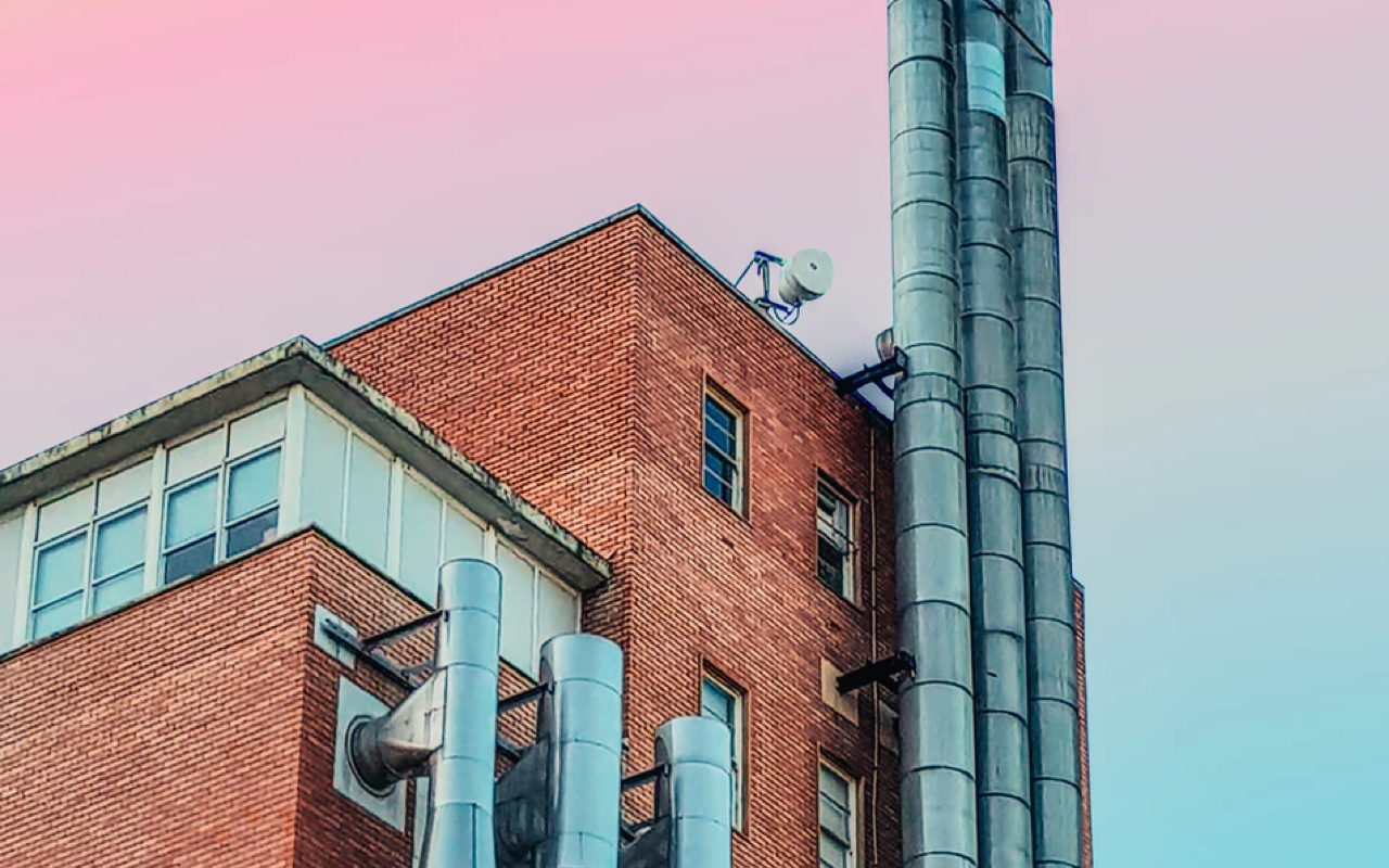 a brick factory looking building with silver tubes on the outside with a bubblegum pink sky in the background